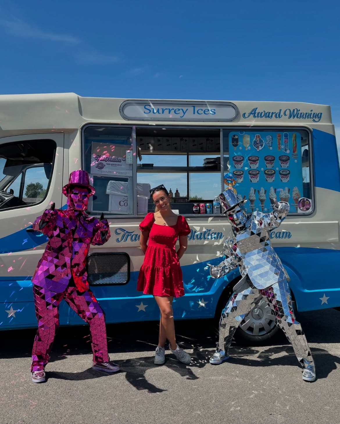 A woman in a red dress standing between two mirrored costumes in front of a blue and cream Hampshire Ices van