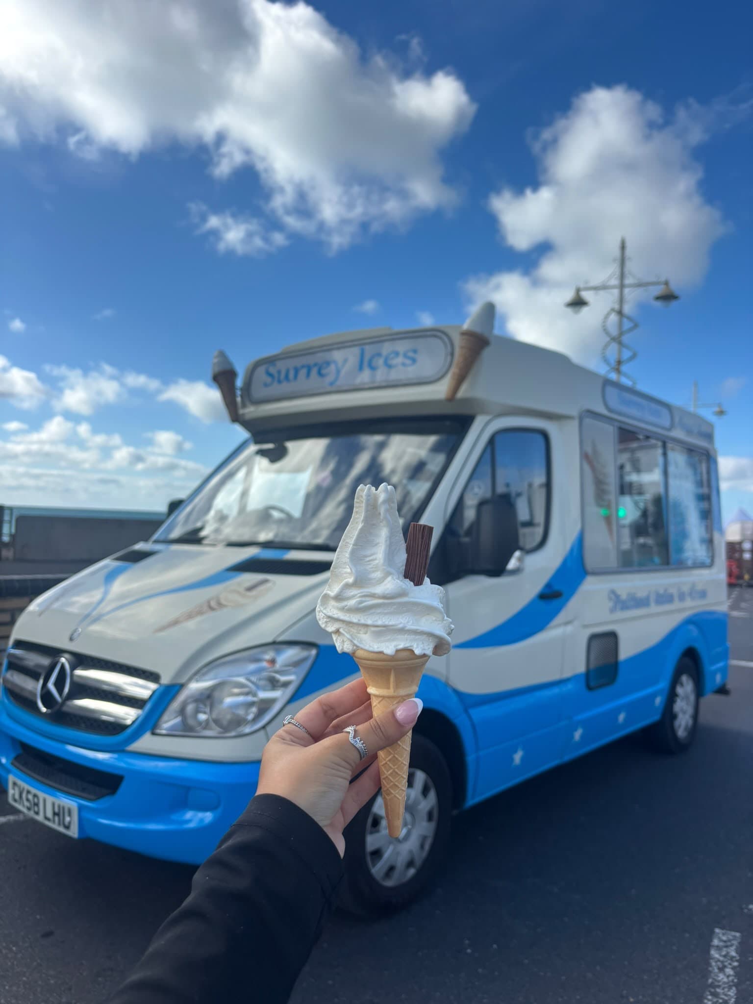 A hand holding a soft serve cone with a chocolate flake in front of a blue and cream Hampshire Ices van