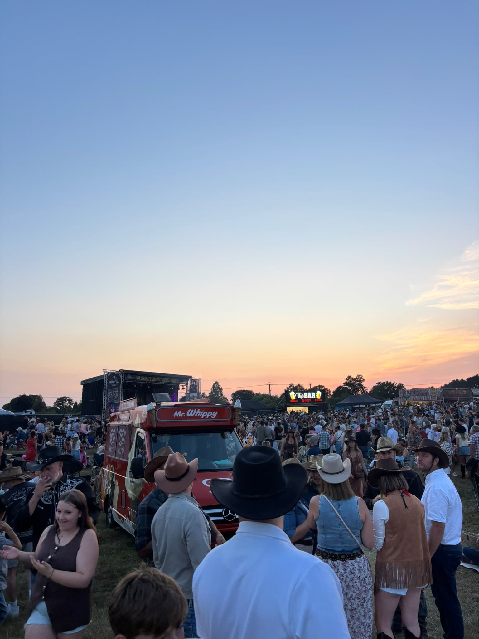 A crowd in hats near a red Mr Whippy van at an outdoor event at dusk, with a stage behind them